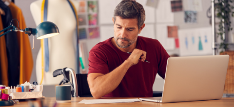 Photo pf a man holding a pen, looking at some paper and working at a laptop.