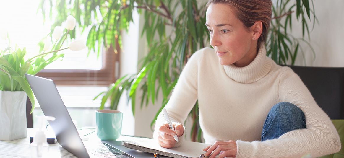 Lady writing on paper whilst looking at laptop